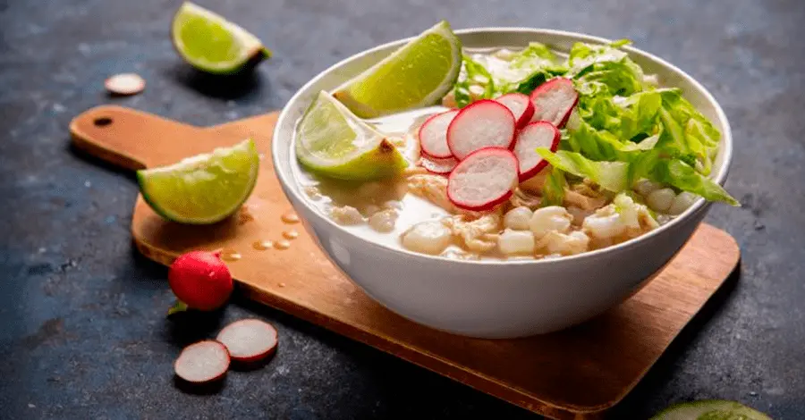 Plato con pozole servido sobre una tabla de cortar con algunos rábanos y limones alrededor