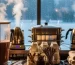 Dispensador transparente sirviendo chocolate caliente espeso con vapor suave; a un lado hervidor y vaporeras en uso; charola con tazas y toppings (canela, ralladura de chocolate); fondo frío con ventana lluviosa que realza la calidez profesional.