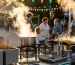 Puesto gastronómico portátil y profesional en una kermés escolar al aire libre durante el atardecer: freidoras con papas burbujeando, plancha portátil en cocción y anafres con olla humeante en primer plano; al fondo banderines de colores, luces cálidas y familias acercándose, transmitiendo profesionalismo, practicidad y sentido de comunidad.